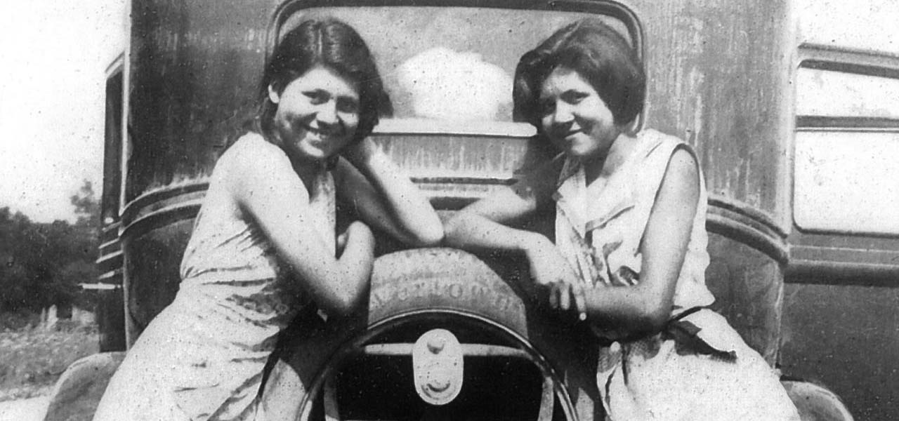Black and white image of two women leaning on an old car.
