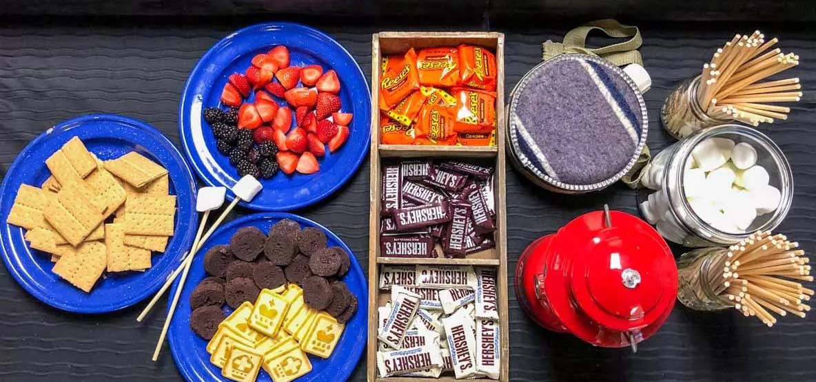 Table spread with items to make s'mores such as marshmallows and chocolate.