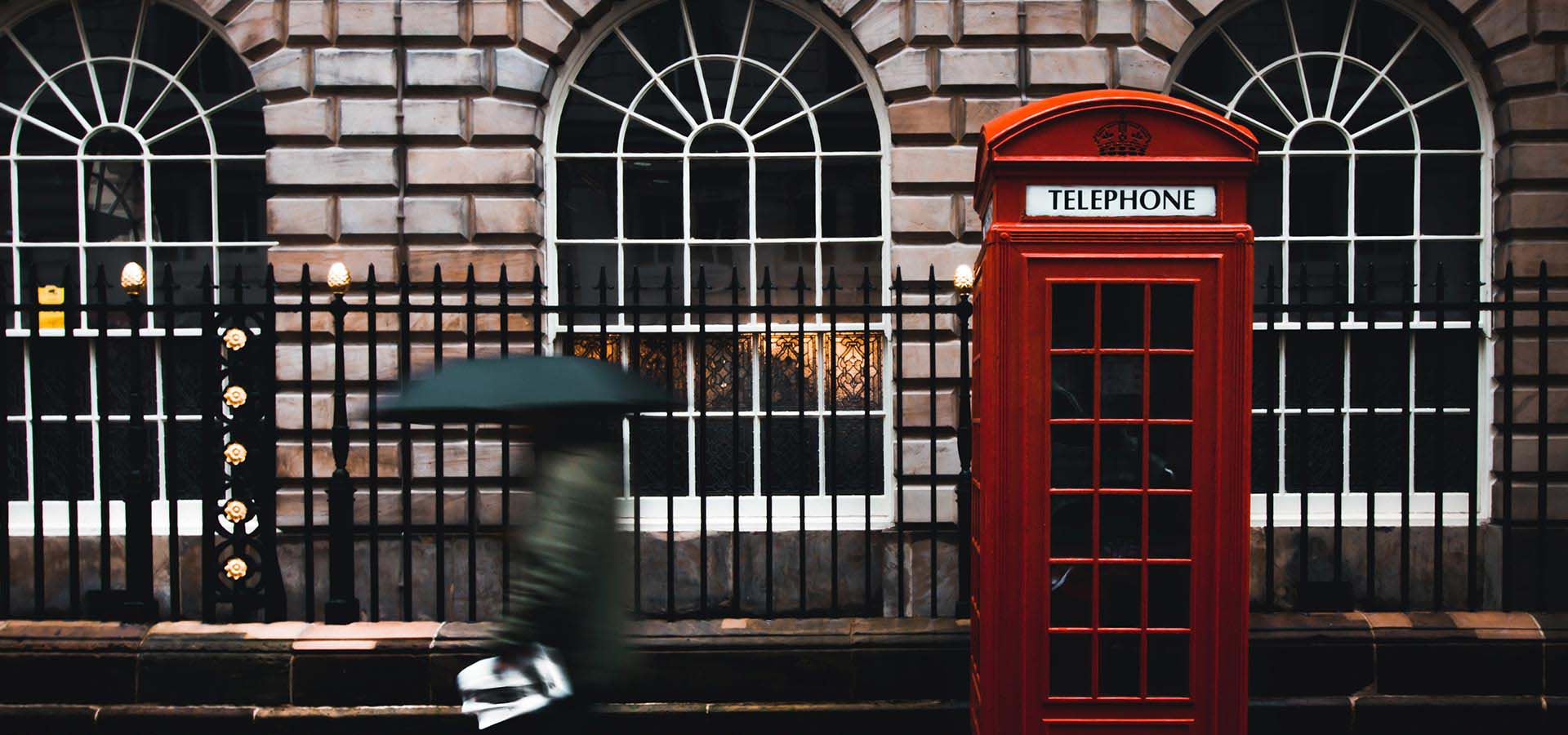 Stone building with red telephone booth in front and a person walking by with a green umbrella.