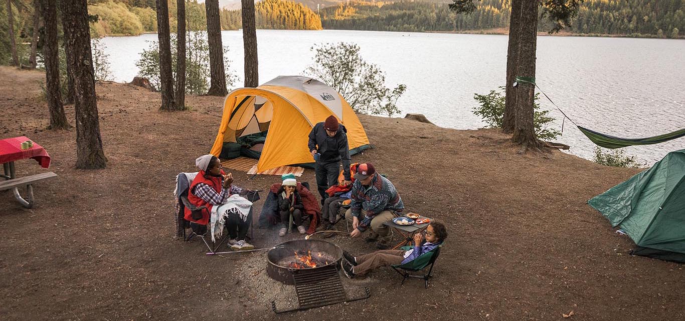 Family around a campfire by an orange tent pitched near a lake.