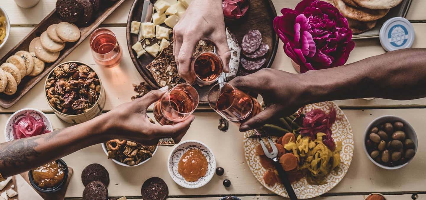 Dining table full of yummy bites of all kinds of foods and 3 people toasting rose wine.