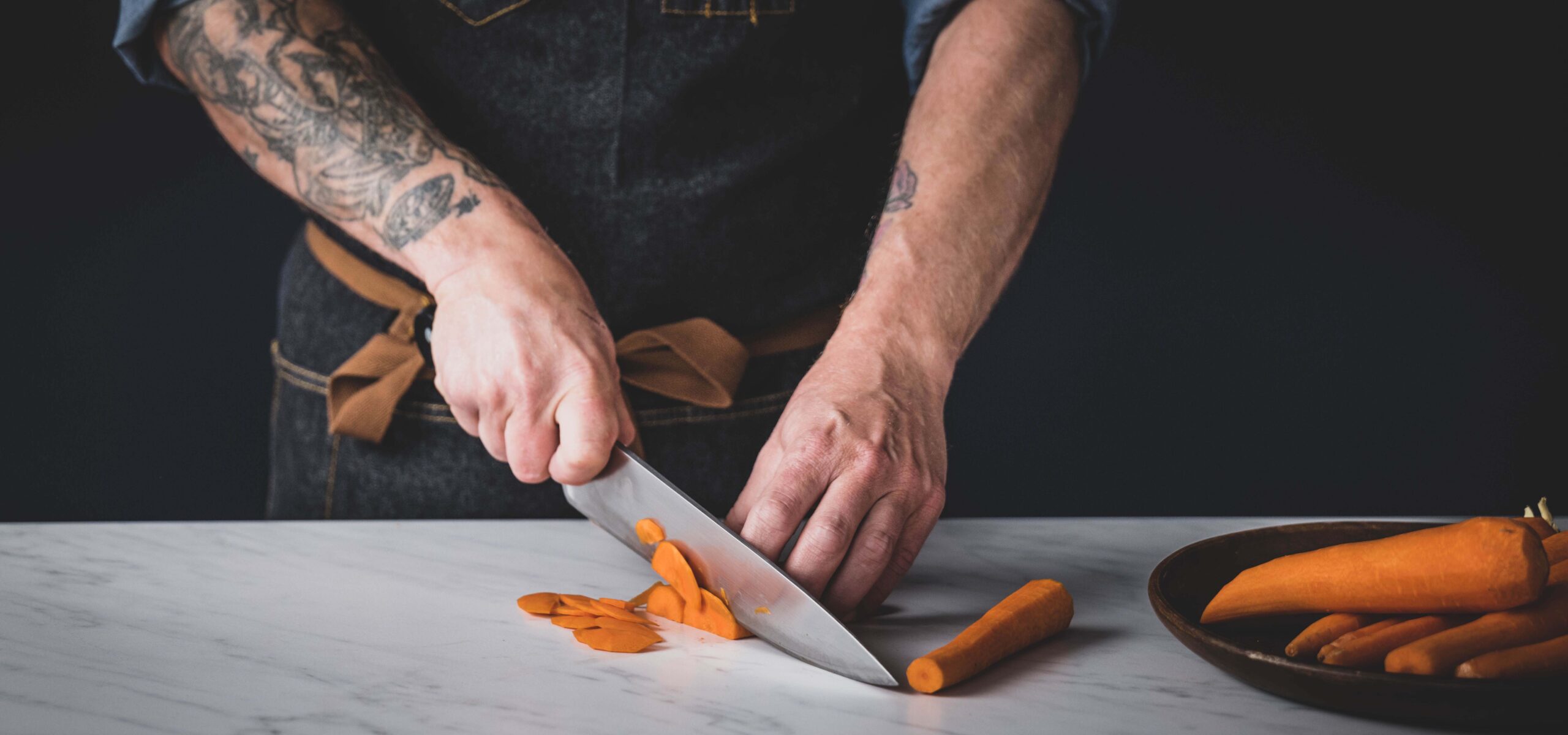 Man with tattooed arms slicing a peeled carrot.