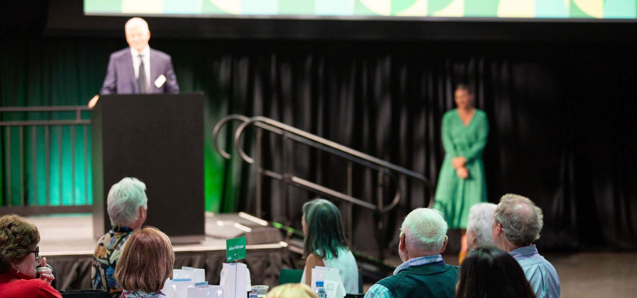 Older man with white hair speaking to a crowd from a stage. A women in a green dress is behind him.