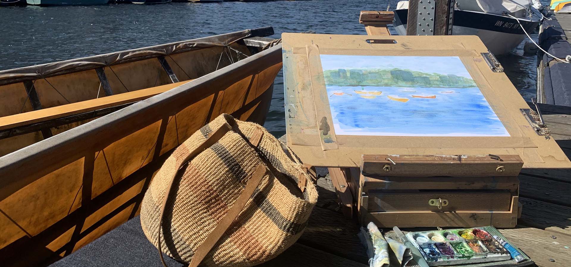 Watercolor of a lake with boats in progress with paint supplies scattered around.