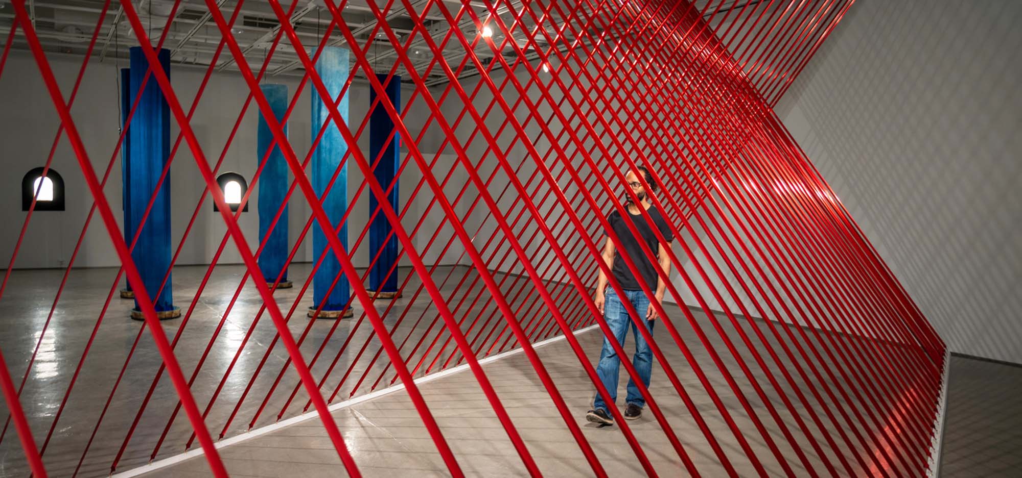 Man walking through a tunnel made with red thin beams inside a large open space.