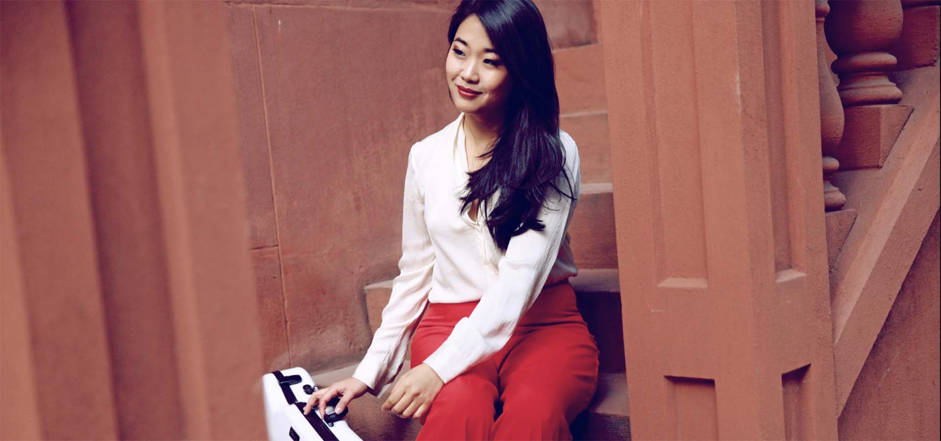 Asian-American woman with white shirt and red pants holding a violin case