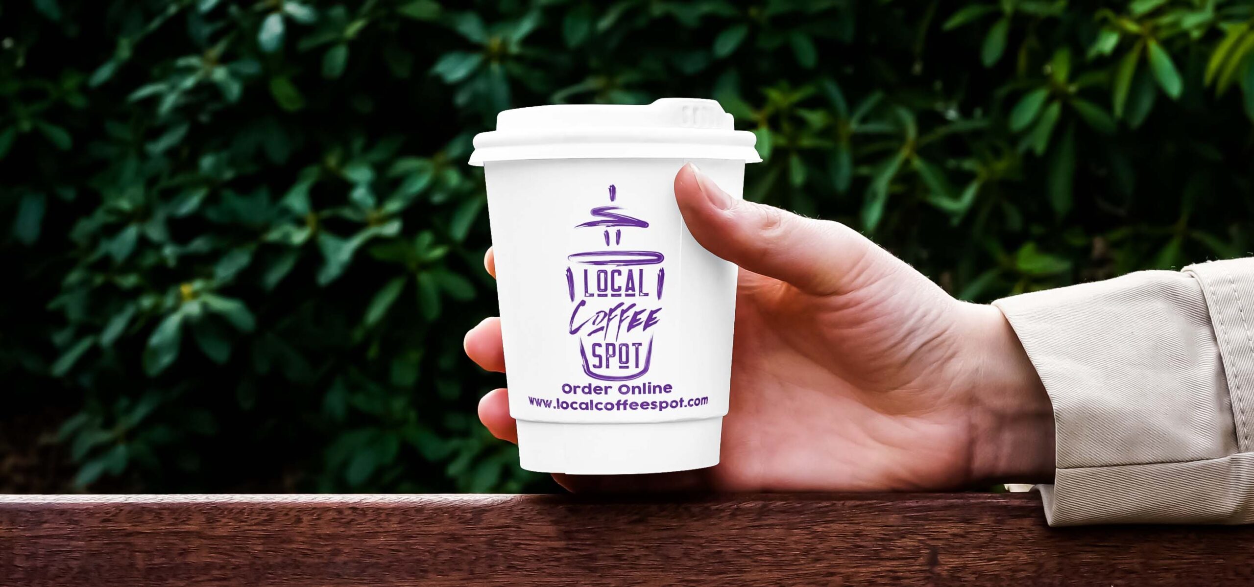 Photo of a hand holding a coffee cup in front of a leafy bush.