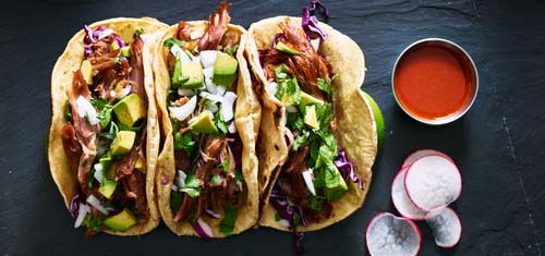 Row of 3 tacos and a side of hot sauce.