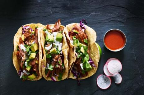 Row of 3 tacos and a side of hot sauce.