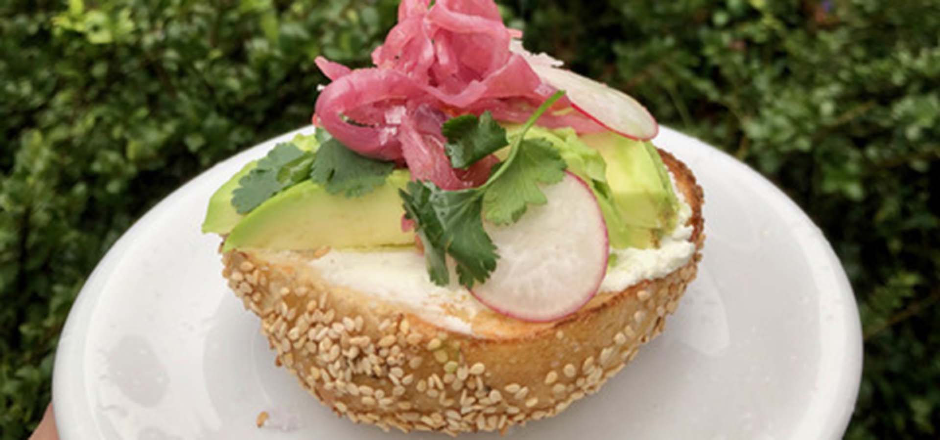 Half a sesame bagel with cream cheese, avocado, radish and pickled onion on a plate.