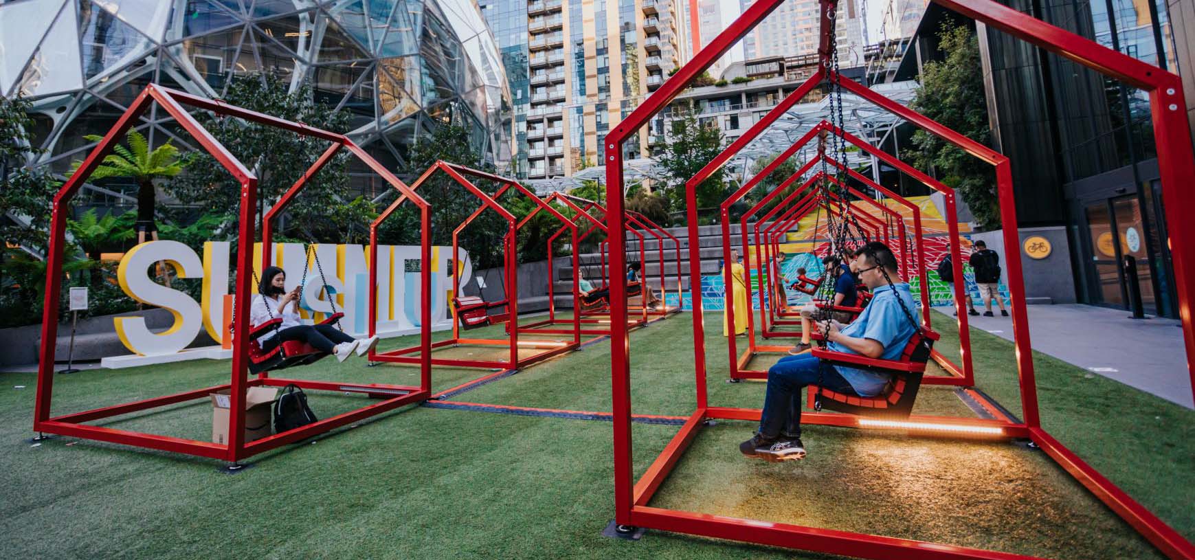 People swinging on outdoor interactive art designed after house porch swings.