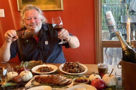 Chef Tom Douglas holding a turkey leg and glass of wine at a Thanksgiving table.