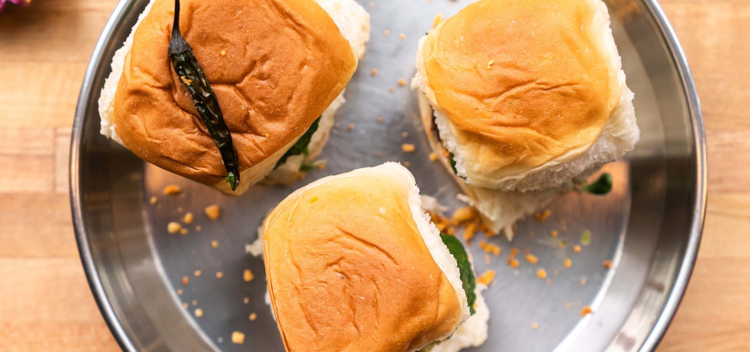 Three Vada Pav sliders on a metal dish; one topped with a roasted green chili pepper.