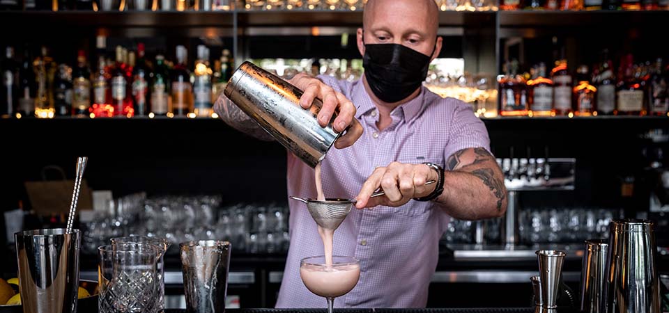 Bartender straining a mixed pink drink into a stem glass.