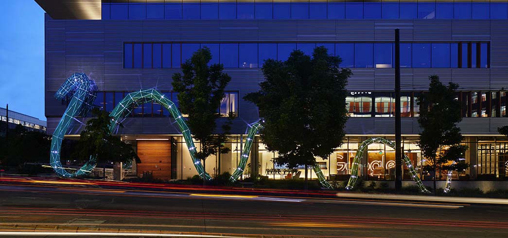 Photo at dusk of a snake-like illuminated outdoor sculpture near an office building.