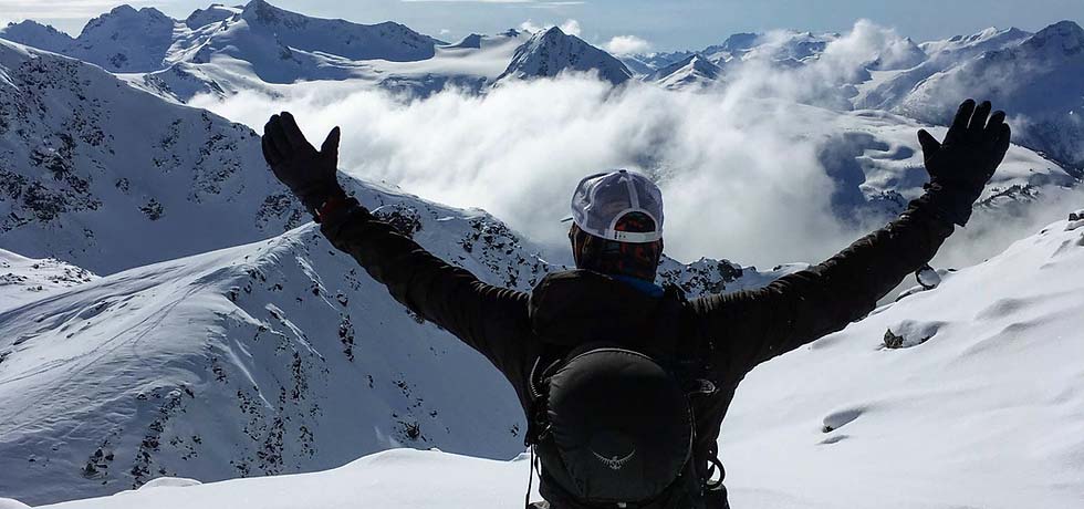 Man at the top of a snowy peak looking at the view with his arms up in amazement.