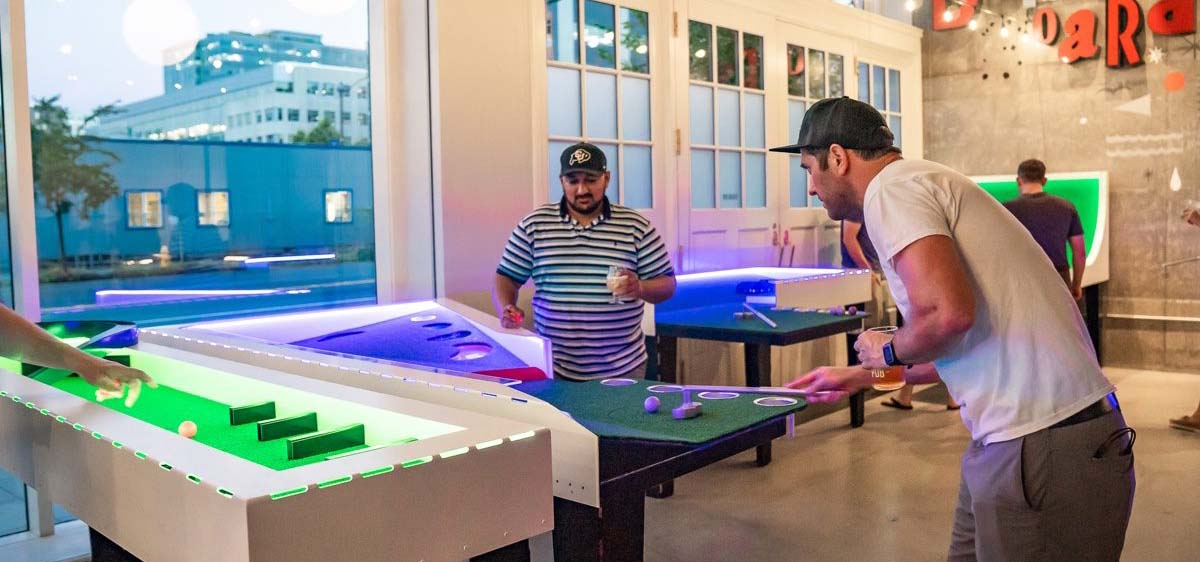 Two men drinking beers and playing tabletop shuffleboard games.